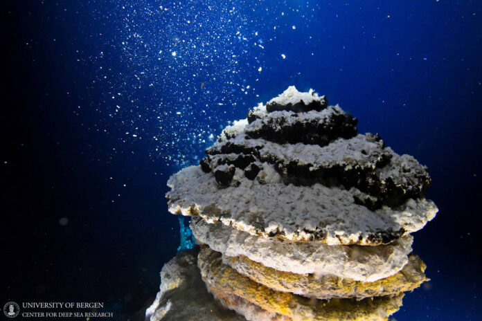 A imagem mostra uma estrutura subaquática que se assemelha a uma rocha ou um coral, com várias camadas e texturas. Bolhas de ar estão subindo ao redor da estrutura, e o fundo é de um azul profundo, típico de ambientes marinhos. A iluminação destaca os detalhes da superfície da estrutura, enquanto a água ao redor parece clara e tranquila.