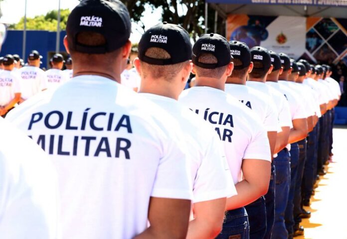 Policiais militares usam camisa branca com o nome da corporação em preto e bonés. Eles estão em fila e de costas
