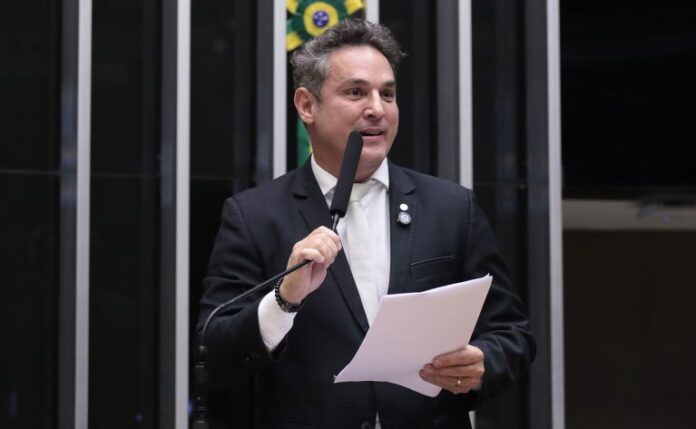 Deputado Zucco fala ao microfone. Ele é um homem claro, com cabelo curto e escuro. Veste um terno azul-marinho e segura um papel