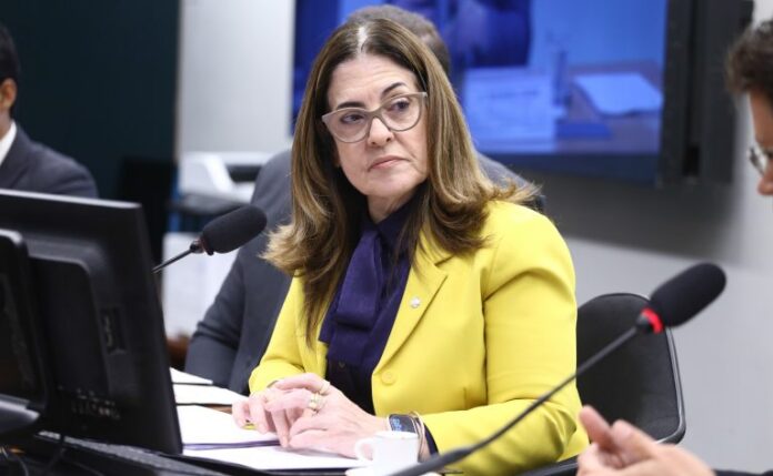 Deputada Rogéria Santos fala ao microfone. Ela é uma mulher clara, com cabelo castanho médio, usa óculos e blazer amarelo e uma camisa azul-marinho 