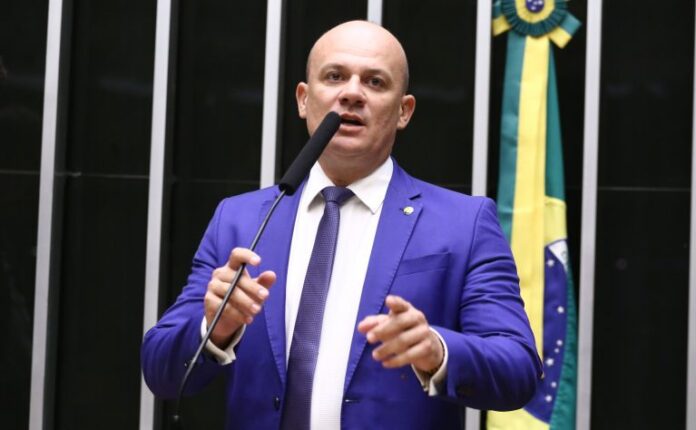 Deputado Cabo Gilberto Silva fala ao microfone. Ele é um homem claro, calvo, usa terno azul marinho e camisa branca. Ao fundo, no lado direito, há uma bandeira do Brasil